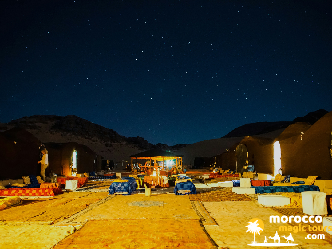A premium mobile camp layout in the Moroccan desert lit by warm lanterns under a clear sky full of stars.