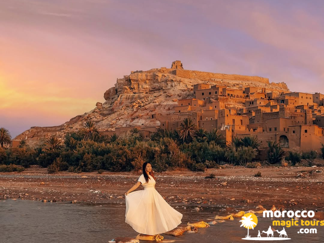 A traveler in a long white dress stands on stones in a riverbed looking up at the Ait Benhaddou Kasbah during a golden sunset.