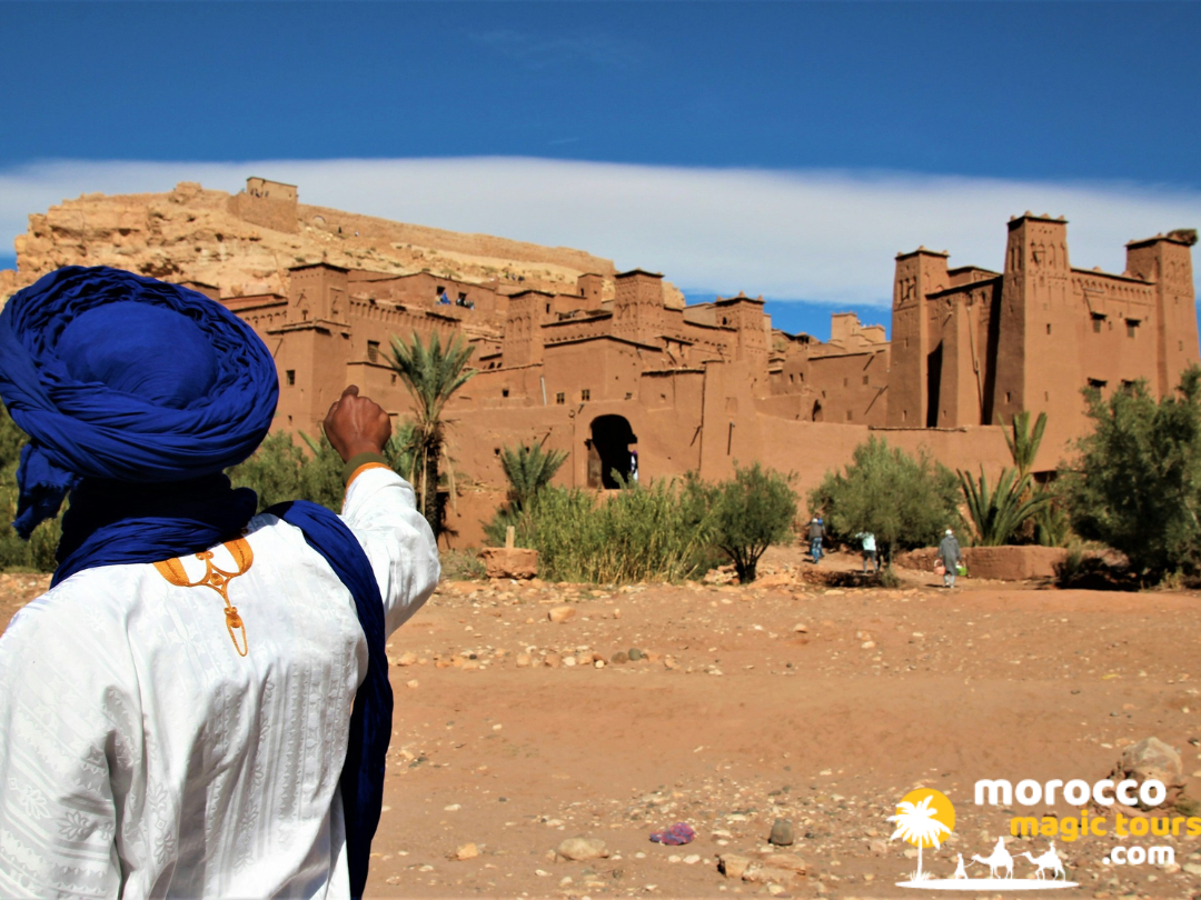 A local guide wearing a traditional blue turban points toward the historic earth fortress of Ait Benhaddou during a 2 day desert tour Marrakech.