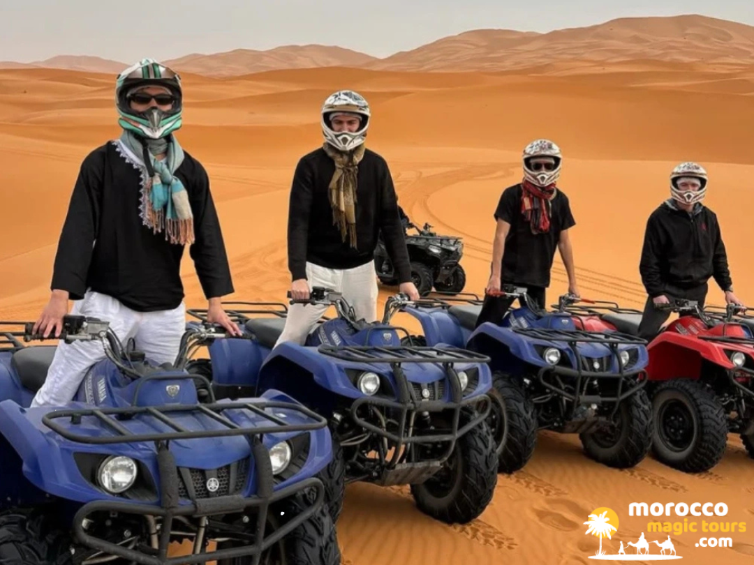 Four friends wearing safety helmets and scarfs posing on quad bikes over rolling desert sands during a Morocco Sahara desert tour.