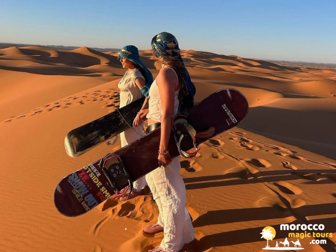Two travelers wearing traditional nomadic headscarves holding sandboards on top of a desert ridge during a 5 Days Tour from Fes to Marrakech excursion.