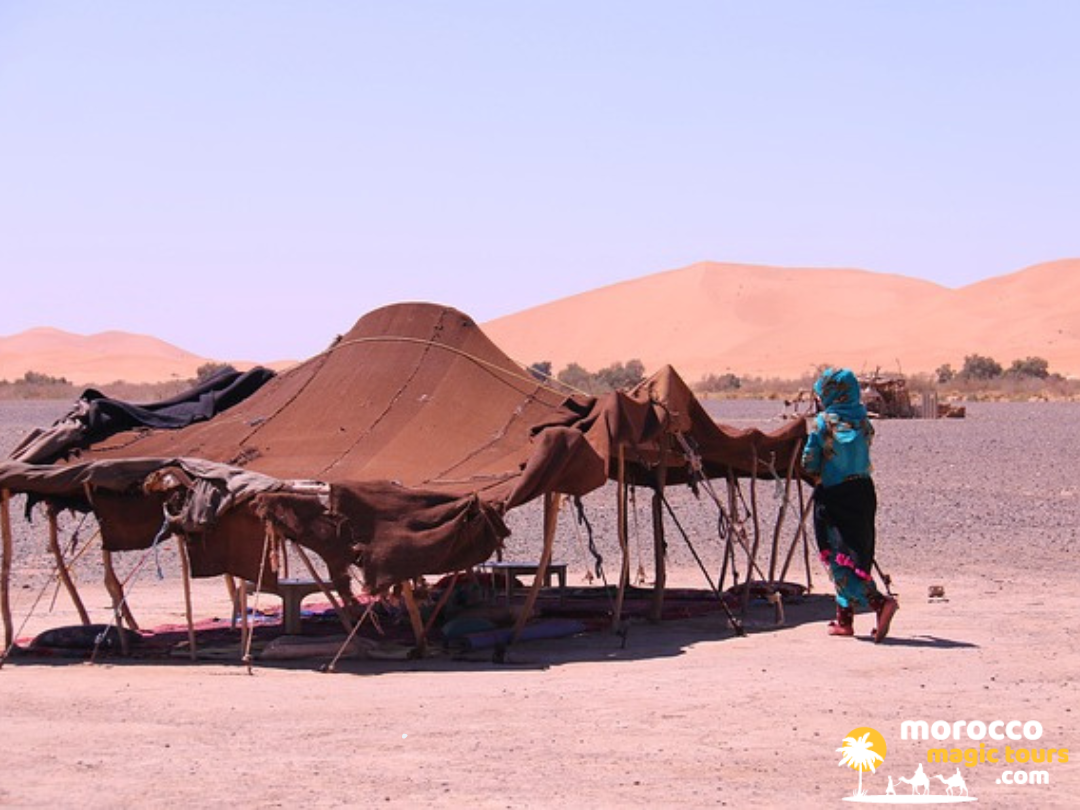A traditional dark woven nomad tent pitched on rocky terrain with sweeping sand dunes in the background during a 5 day tour Morocco.