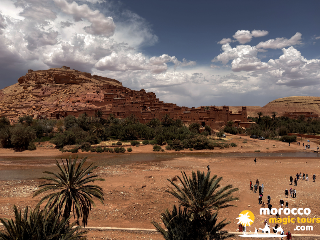 Wide panoramic view of the UNESCO World Heritage site Ksar Aït Benhaddou with palm trees in the foreground.