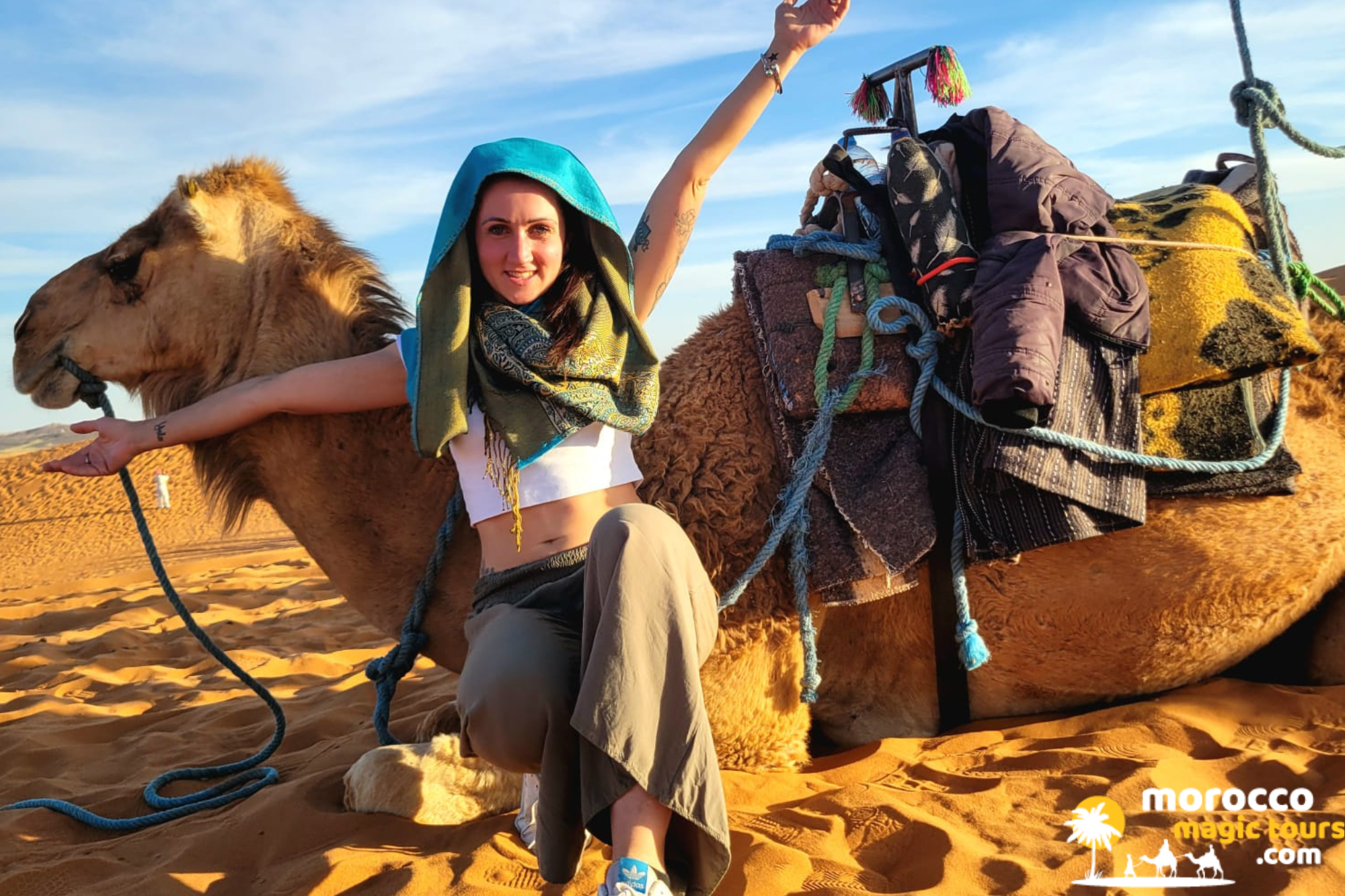 Traveler smiling next to a saddled dromedary in the golden Merzouga sand dunes on a desert tour from Tangier