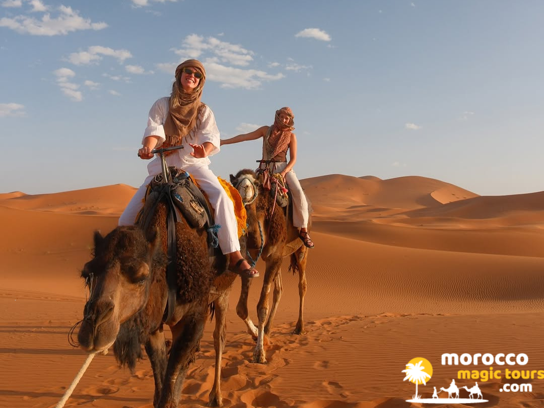Two happy female tourists in headscarves riding dromedaries over bright orange desert dunes on a 5 day desert tour from marrakech