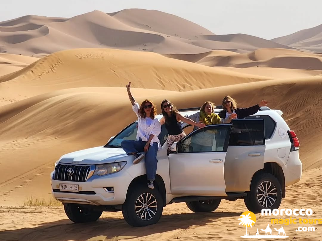 A group of happy female tourists posing on a luxury white 4x4 Toyota SUV parked among the sand dunes of Erg Chebbi during a 5 day desert tour from marrakech.