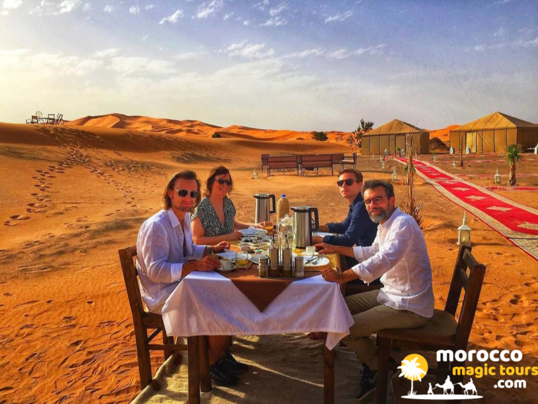 Four friends having breakfast on a table outside on a red carpet in a desert camp on a 5 day desert tour from marrakech.