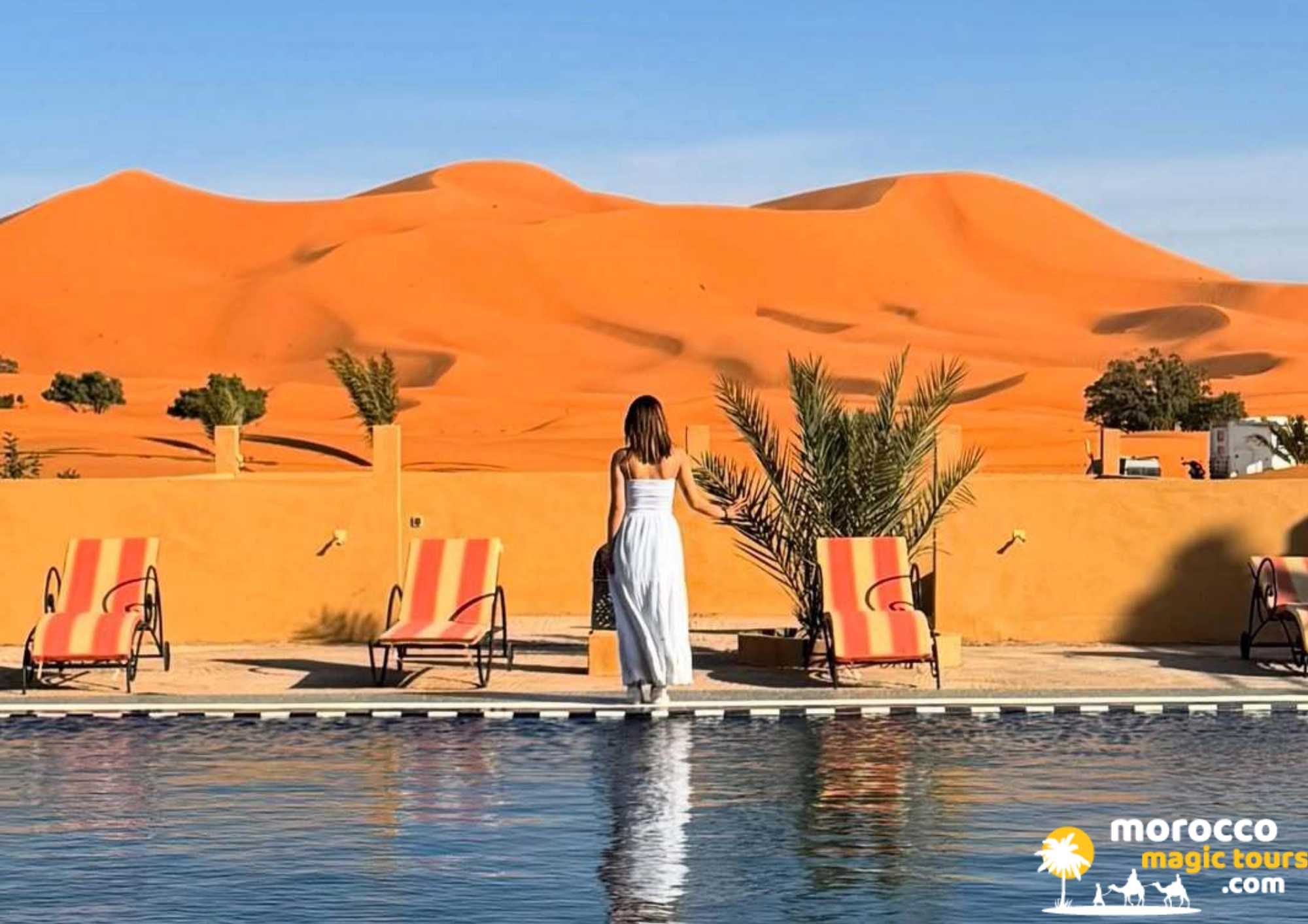 A woman in a white dress stands by a clear blue swimming pool with views over the orange dunes of Erg Chebbi in Merzouga, Morocco. 8 Days From Casablanca Sahara Desert Tour - Day 3