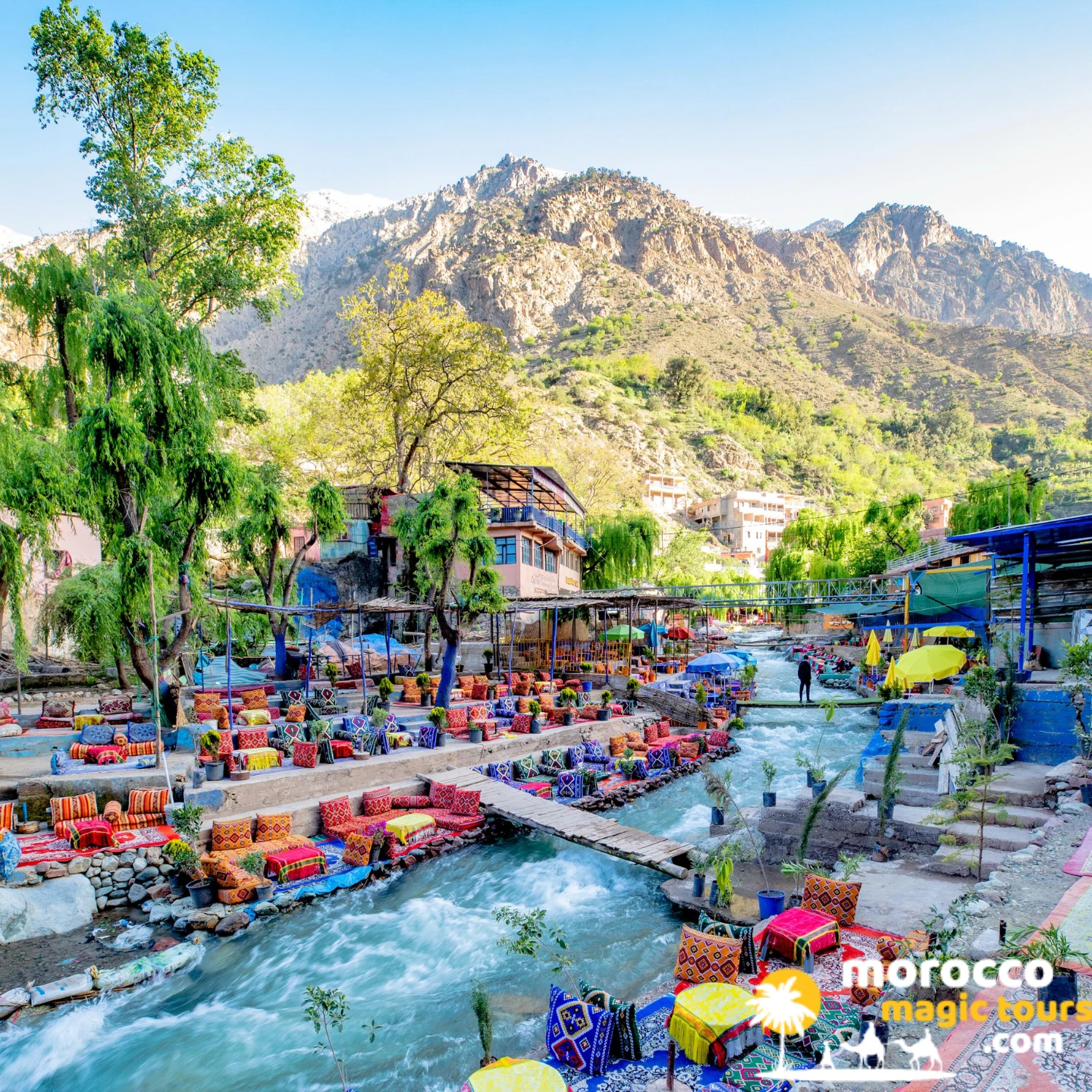Colorful riverside lounge cushions and wooden bridges over the Ourika River with snow-capped High Atlas peaks