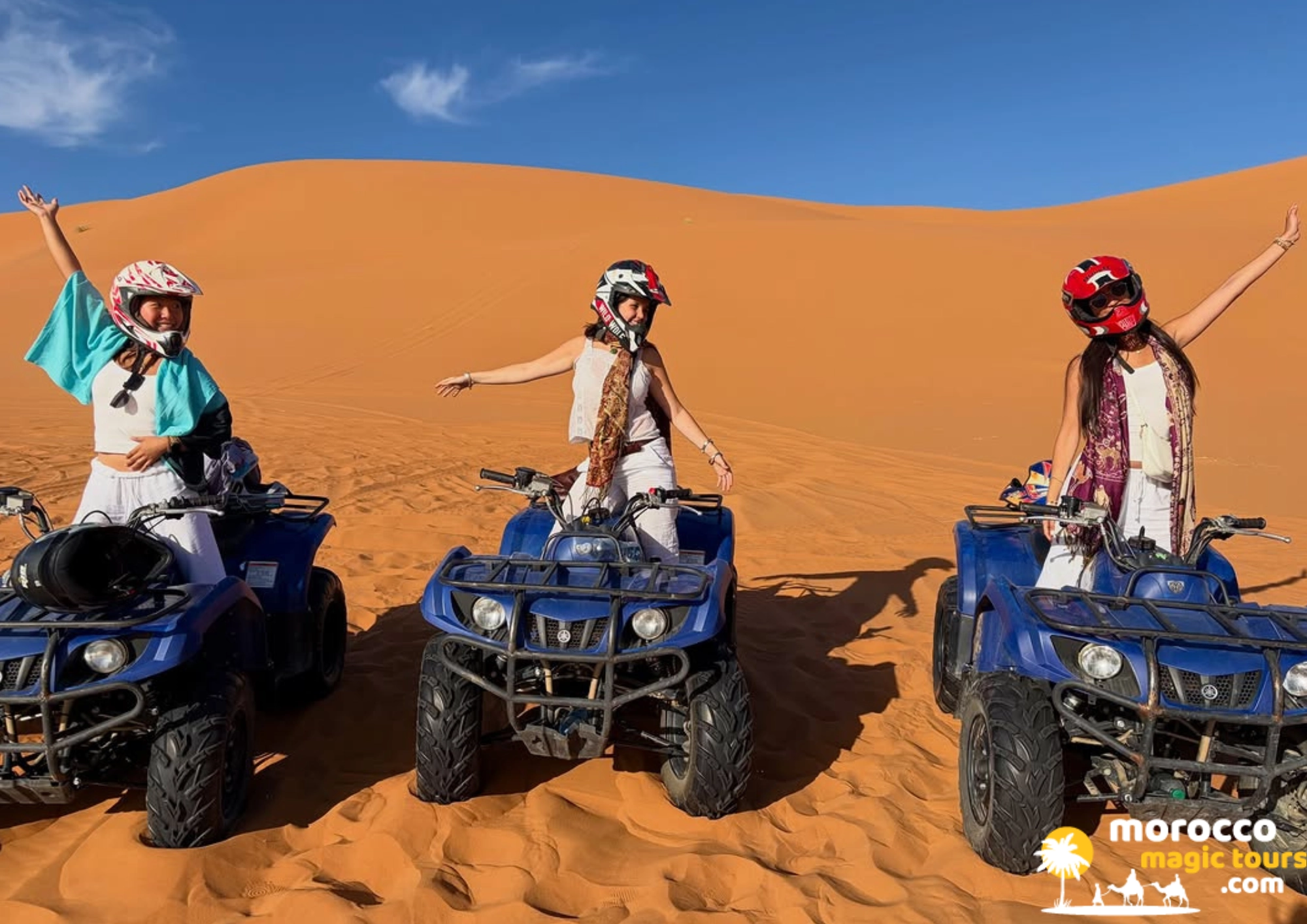 Three happy women riding blue quad bikes in front of a big sand dune during an 8 days tour Morocco.
