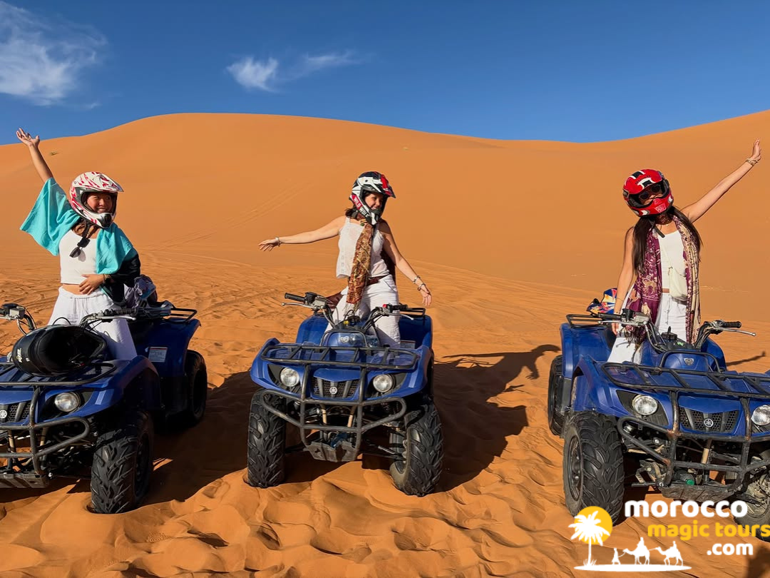 Three smiling women wearing safety helmets pose on blue quad bikes in the orange sand dunes on a thrilling 5 day desert tour from marrakech