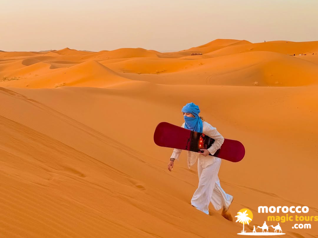 An adventurous traveler wearing a traditional blue headscarf carrying a snowboard up an expansive orange sand dune on a 5 day desert tour from marrakech.