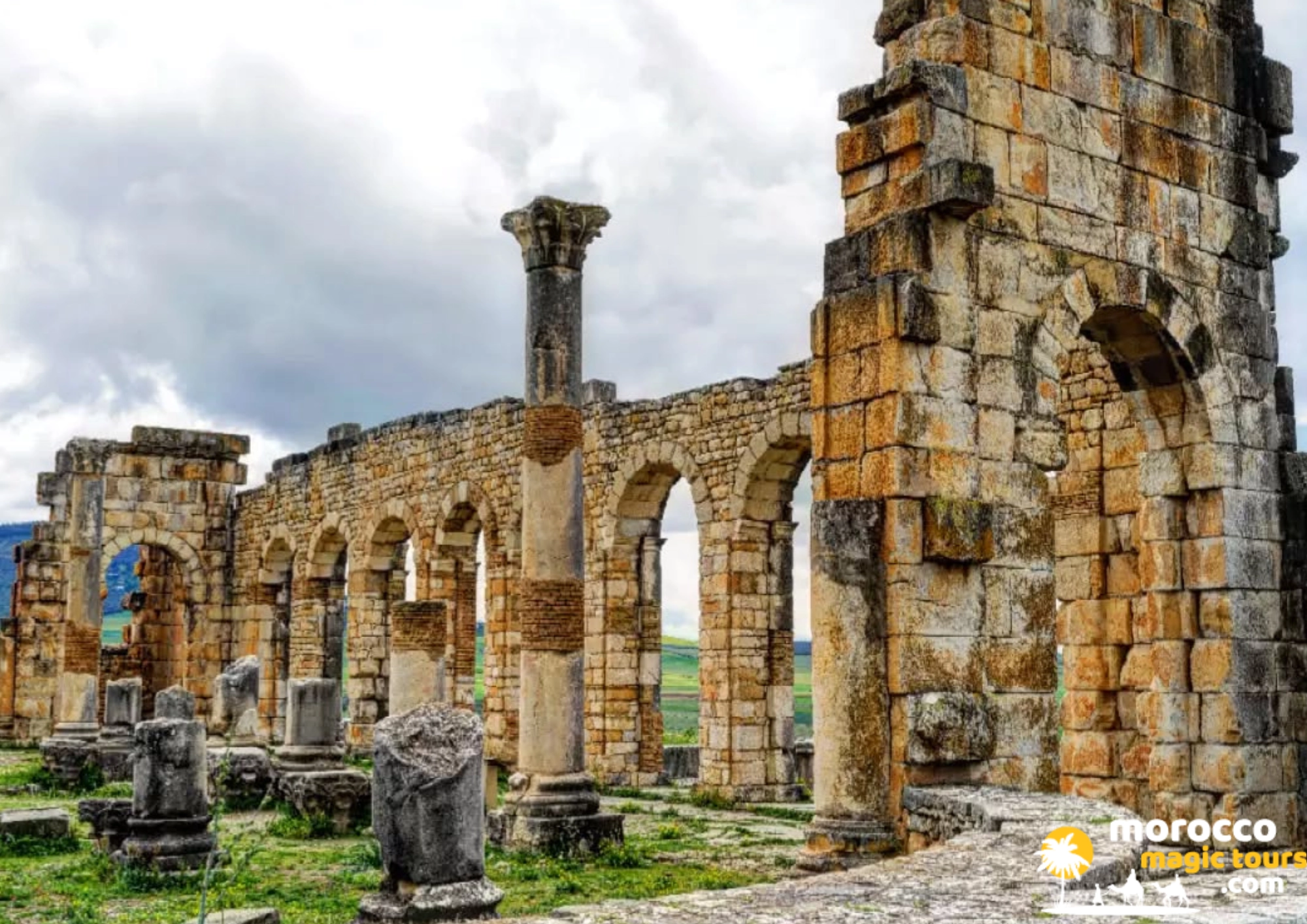 Stone columns and arched ruins of the Roman city of Volubilis, a UNESCO World Heritage Site, under a cloudy sky. during an 8 days casablanca tour.
