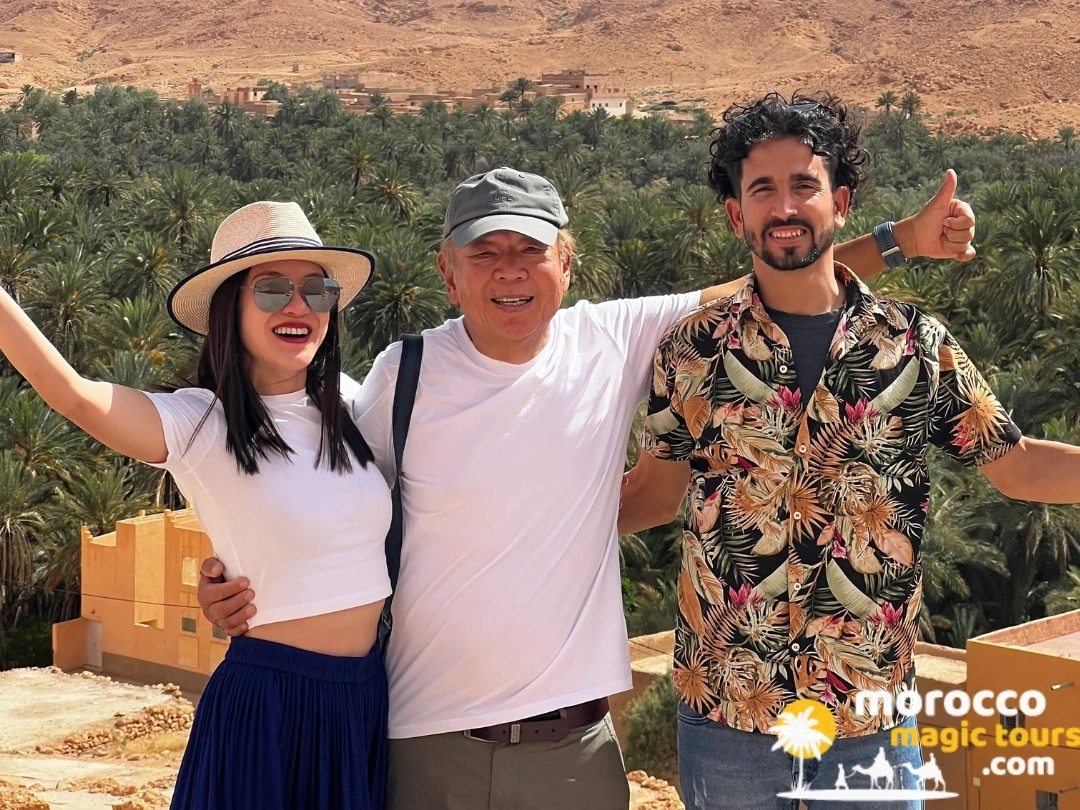 Viajeros que sonríen juntos en un mirador panorámico con vistas a un frondoso palmeral y a las laderas desérticas, de la mano de Morocco Magic Tours.