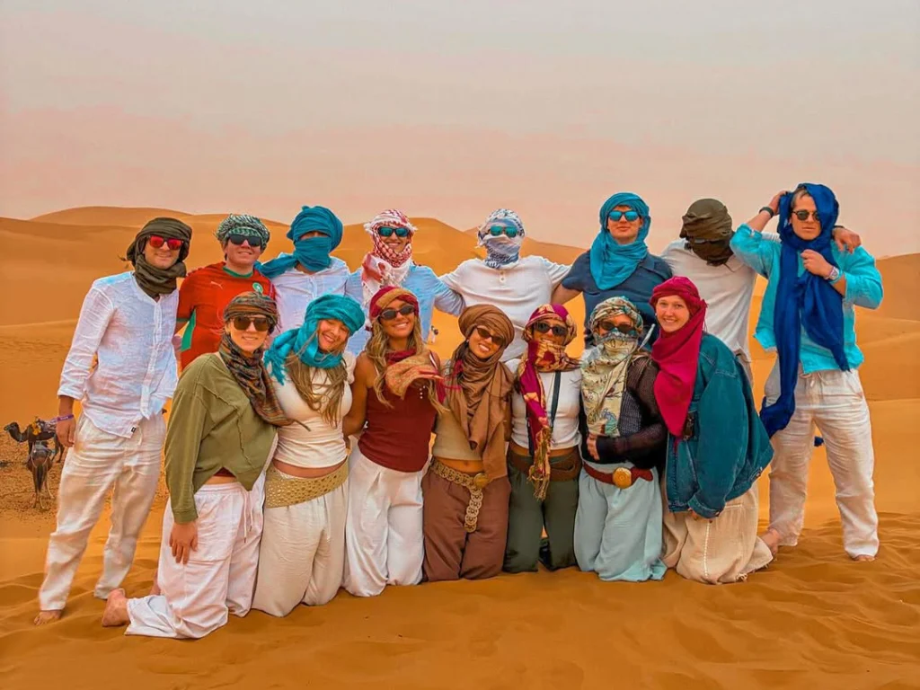 A group of adventurers geared up in quad biking helmets pose on the Merzouga sand dunes, showcasing the thrilling quad-biking excursion available on Morocco Magic Tours' Marrakech to Fes 3-day Sahara Desert Tour.