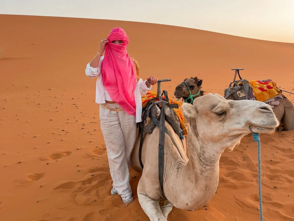 A traveler in a pink headscarf stands beside her camel on the golden dunes at sunset, capturing the iconic camel trek featured on Morocco Magic Tours' 3-day Sahara Desert Tour from MARRAKECH.