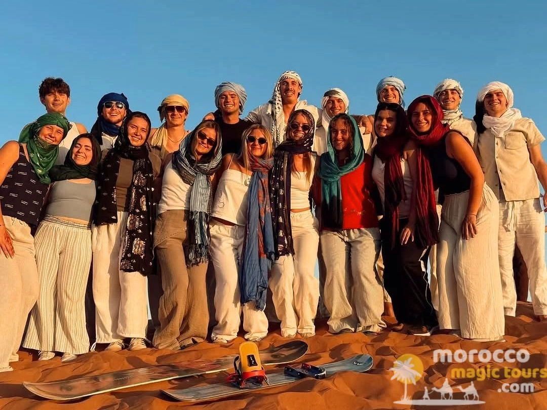 Un gran grupo de jóvenes viajeros estudiantes con pañuelos tradicionales de pie sobre las dunas de arena con tablas de sandboard durante un viaje por el desierto de Morocco Magic Tours.