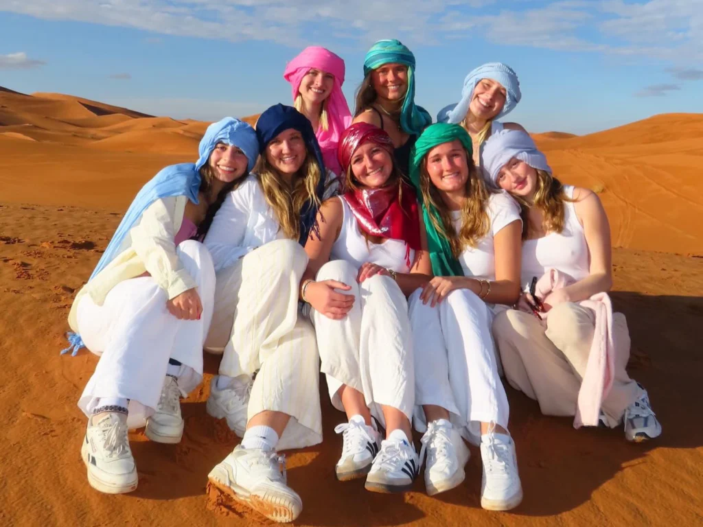 Travelers wearing traditional Berber turbans sit together on the Erg Chebbi dunes at golden hour, a signature Sahara Desert Tour moment offered on Morocco Magic Tours' Marrakech to Merzouga itineraries.