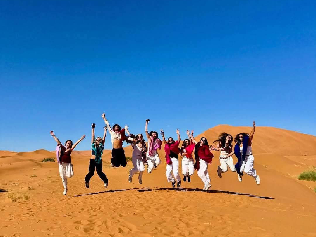 A lively group of travelers jumps together atop the golden Erg Chebbi dunes, capturing the excitement of a Marrakech to Merzouga Sahara Desert Tour with Morocco Magic Tours.